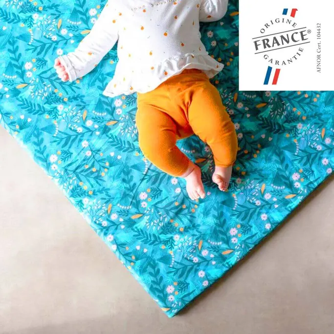 Un bébé en pantalon orange assis sur un tapis d'éveil turquoise à fleurs, avec le logo Origine France Garantie en coin