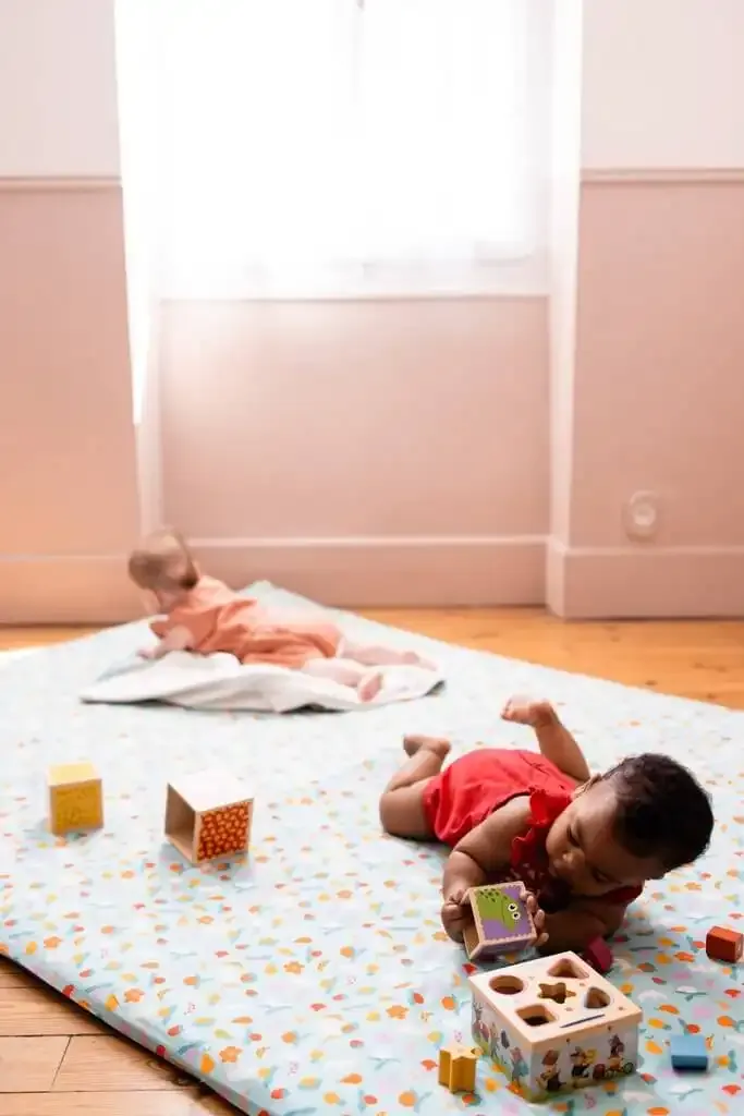 Deux bébés sur un grand tapis d'éveil blanc à petits motifs posé sur un parquet, l'un allongé au fond, l'autre qui joue avec des cubes en bois