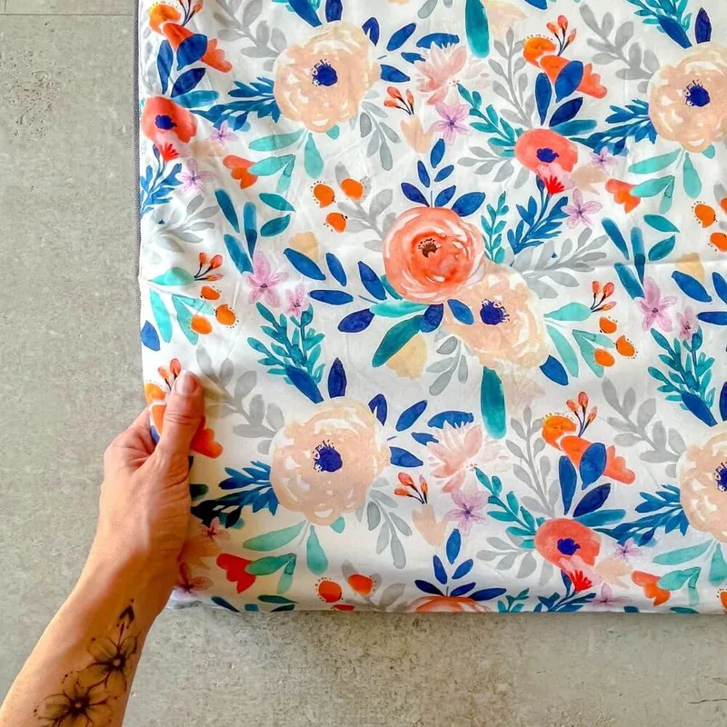 Une main qui soulève le coin d'un tapis d'éveil blanc imprimé de fleurs orange et bleues