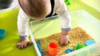 Un bébé penché sur un bac transparent rempli de graines, avec des petits pots et gobelets colorés