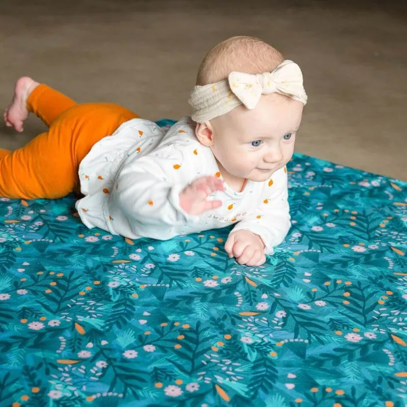 Un bébé avec un bandeau à nœud, allongé sur le ventre sur un tapis d'éveil bleu canard imprimé de feuillages, qui relève la tête
