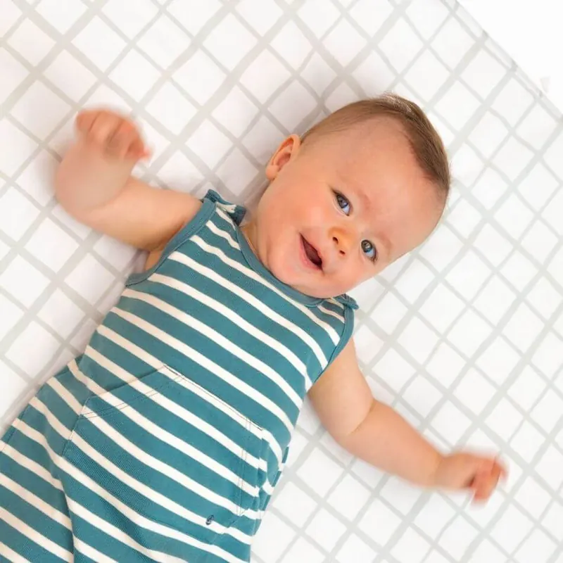 Un bébé souriant en body rayé bleu canard et blanc, allongé sur le dos sur un tapis d'éveil blanc à carreaux gris : découvrir le tapis magique