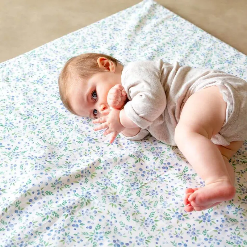 Un bébé allongé sur le côté sur un tapis d'éveil blanc imprimé de petites fleurs bleues, la main près de la bouche