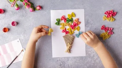 Des mains d'enfant qui collent des pâtes papillon colorées en forme de bouquet sur une feuille blanche