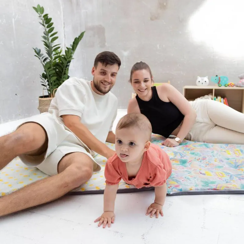 Un bébé à quatre pattes sur un tapis d'éveil imprimé, entre son père et sa mère assis au sol