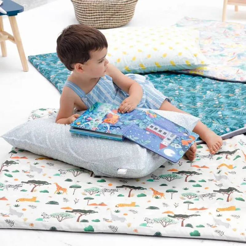Un petit garçon allongé sur des coussins, qui feuillette un livre sur des tapis d'éveil imprimés posés au sol
