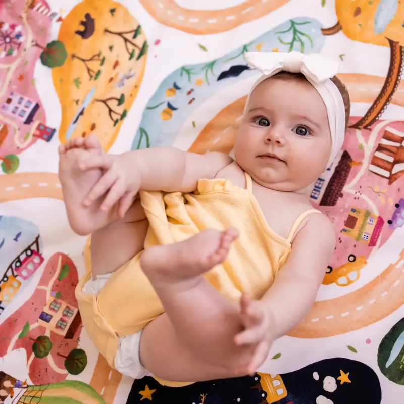 Un bébé avec un bandeau blanc, allongé sur le dos sur un tapis d'éveil imprimé de maisons et de routes colorées, qui attrape ses pieds
