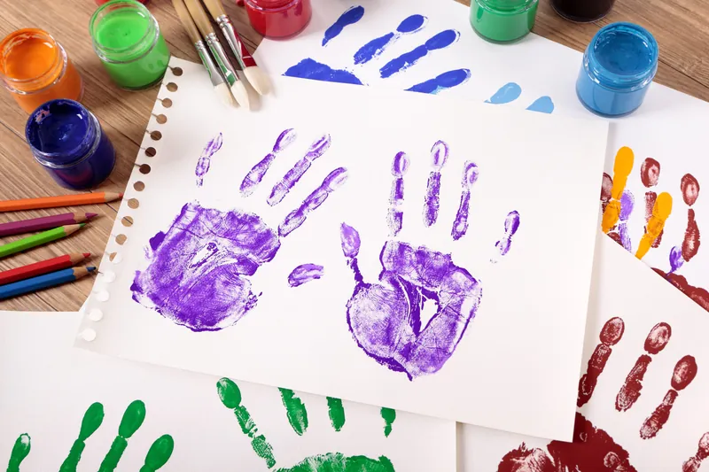 Des empreintes de mains d'enfant à la peinture violette, verte et rouge sur des feuilles blanches, à côté de pots de peinture et de crayons de couleur
