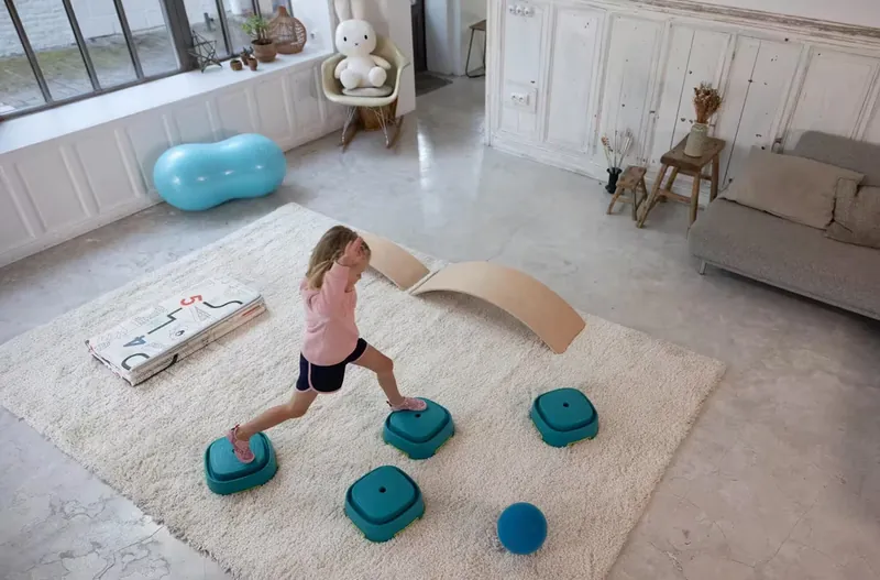 Une petite fille qui enjambe des plots de motricité bleu canard sur un tapis blanc, dans un grand salon lumineux