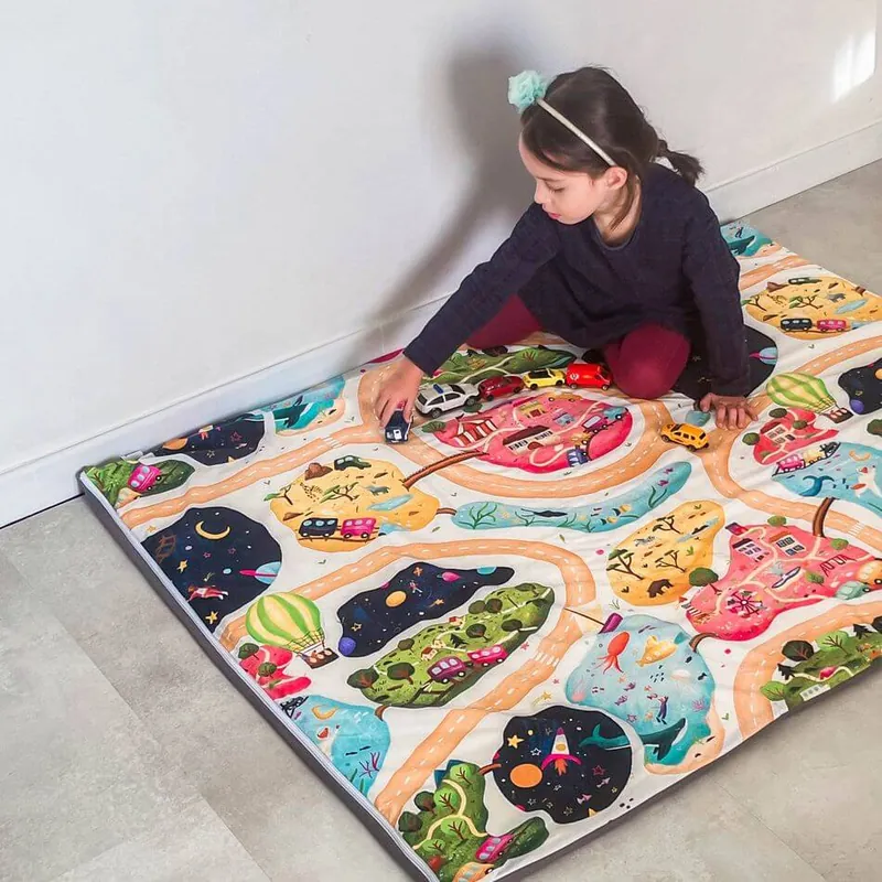 Une petite fille assise sur un tapis d'éveil imprimé de routes et de villages colorés, qui fait rouler une petite voiture