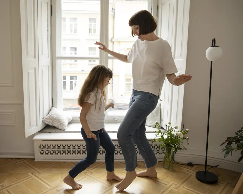 Une mère et sa fille qui dansent pieds nus dans un salon, devant une grande fenêtre