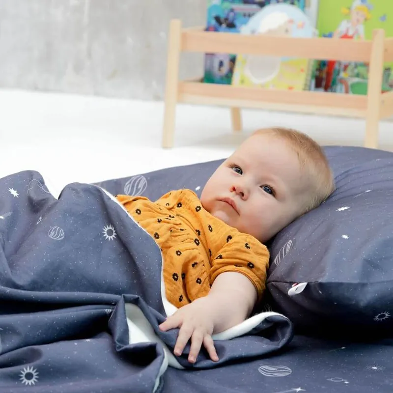Un bébé en haut moutarde allongé sur un oreiller bleu nuit imprimé de planètes, en partie couvert d'une couverture assortie