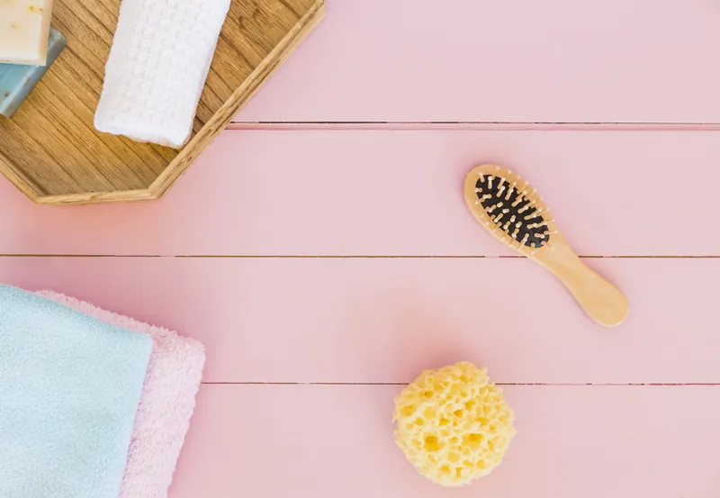 Vue de dessus d'accessoires de toilette pour bébé sur un fond rose : brosse à cheveux en bois, éponge naturelle, serviettes pliées et savon