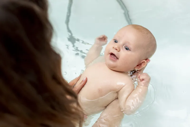 Un bébé souriant dans son bain, soutenu par sa mère dont on voit la chevelure au premier plan