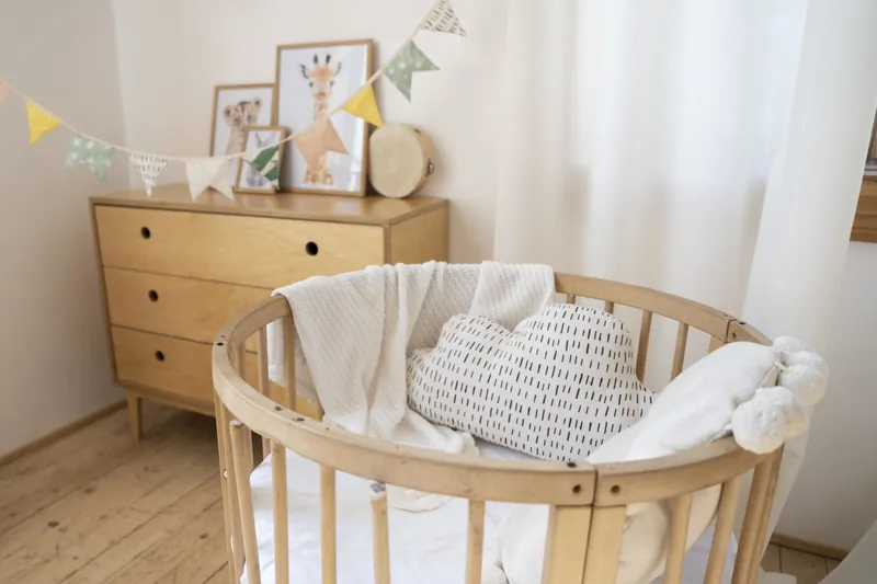 Une chambre de bébé claire : berceau rond en bois garni de coussins, commode en bois, cadres et guirlande de fanions au mur