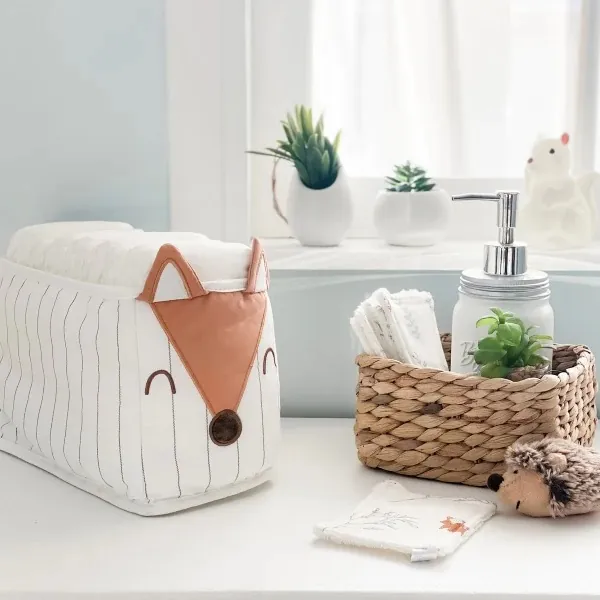 Une panière à couches en tissu blanc à tête de renard, à côté d'un panier en osier garni de lingettes et d'un flacon pompe, devant une fenêtre