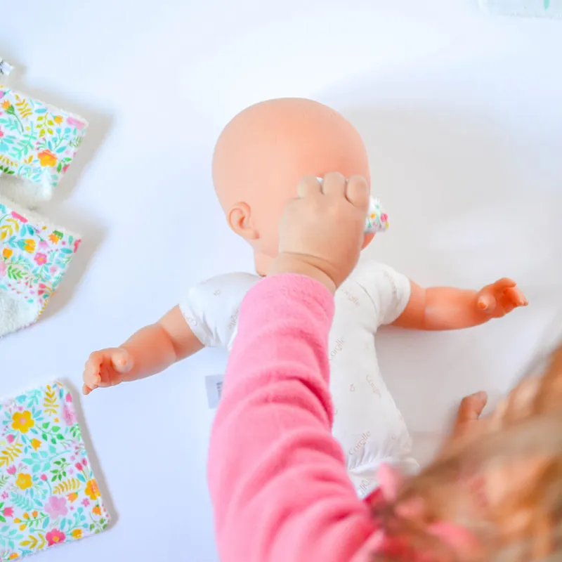 Une enfant en pull rose penchée sur un poupon allongé, à côté de couches et de lingettes en tissu fleuri