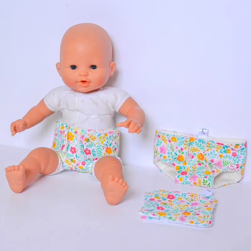 Un poupon assis qui porte une couche en tissu fleuri, une deuxième couche et une lingette posées à côté de lui