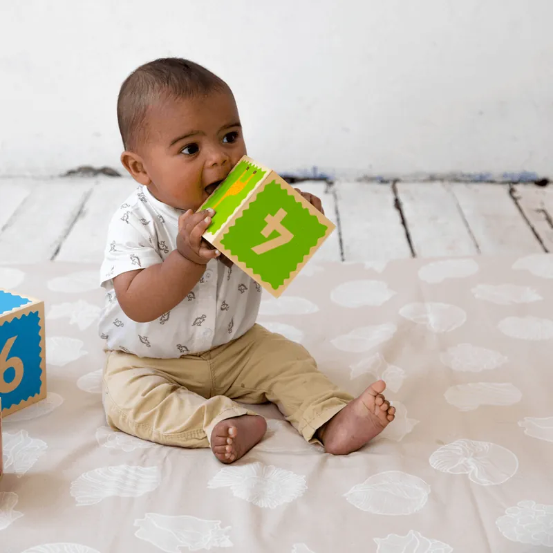 Un bébé assis sur un tapis d'éveil beige imprimé de feuilles, qui mordille un cube en carton vert