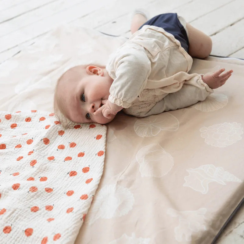 Un bébé allongé sur le côté qui suce son pouce, sur un tapis d'éveil beige posé à côté d'un tapis à pois rouges