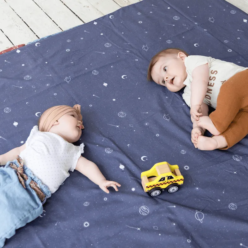 Deux bébés allongés sur un grand tapis d'éveil bleu nuit imprimé de planètes et d'étoiles, une petite voiture jaune entre eux