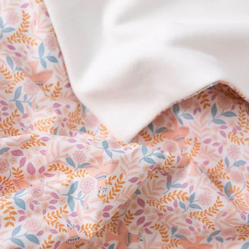 Gros plan sur une couverture bébé en coton imprimé de fleurs et de feuillages orange, roses et bleus, la doublure blanche repliée sur un coin