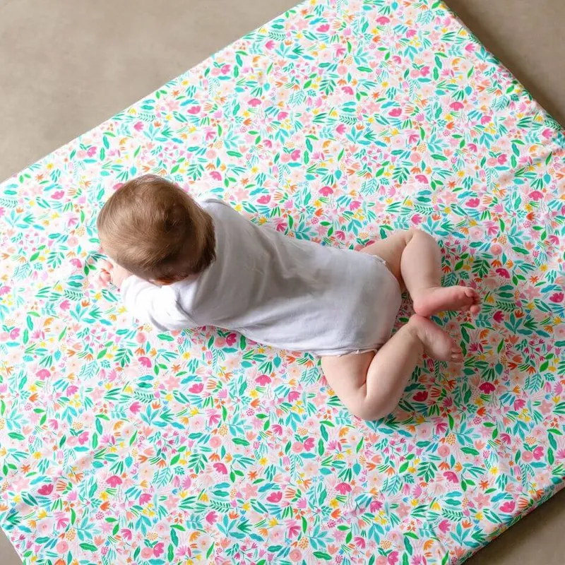 Un bébé en body blanc, vu de dos, qui rampe à plat ventre sur un tapis d'éveil imprimé de fleurs roses, vertes et bleues