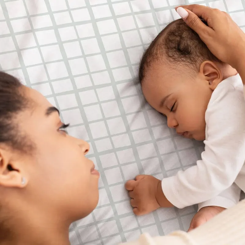 Un bébé endormi sur un tapis d'éveil blanc à carreaux gris, sa mère allongée face à lui qui lui caresse la tête