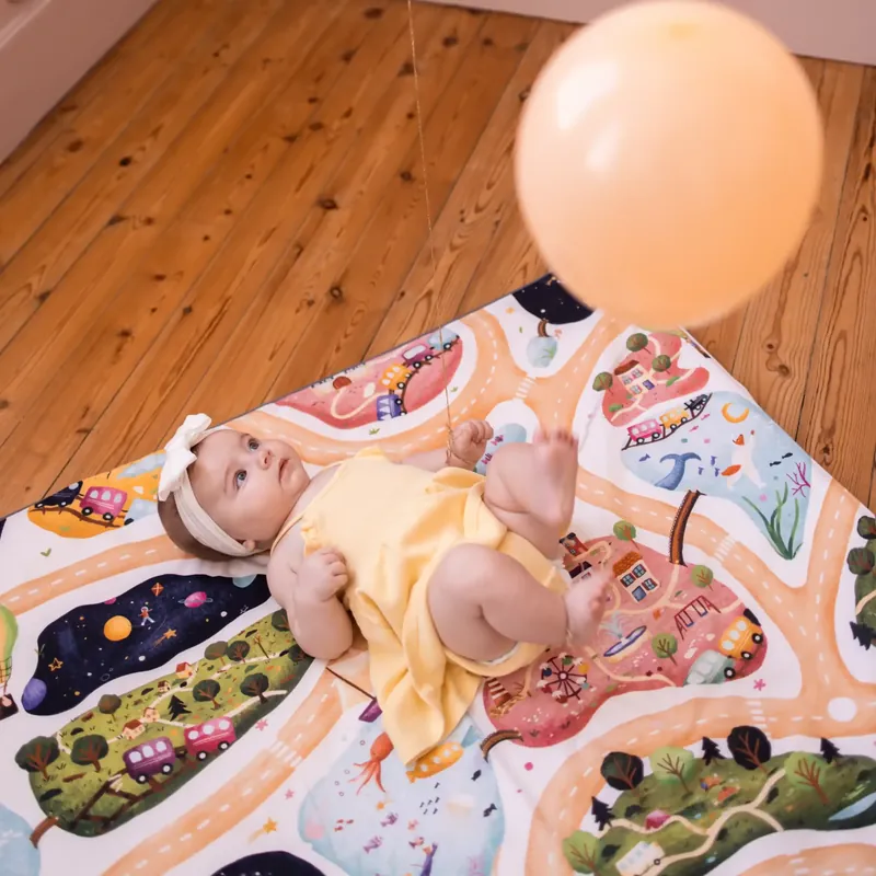 Un bébé en barboteuse jaune et bandeau blanc, allongé sur le dos sur un tapis d'éveil illustré de routes et de paysages, regarde un ballon orange pâle