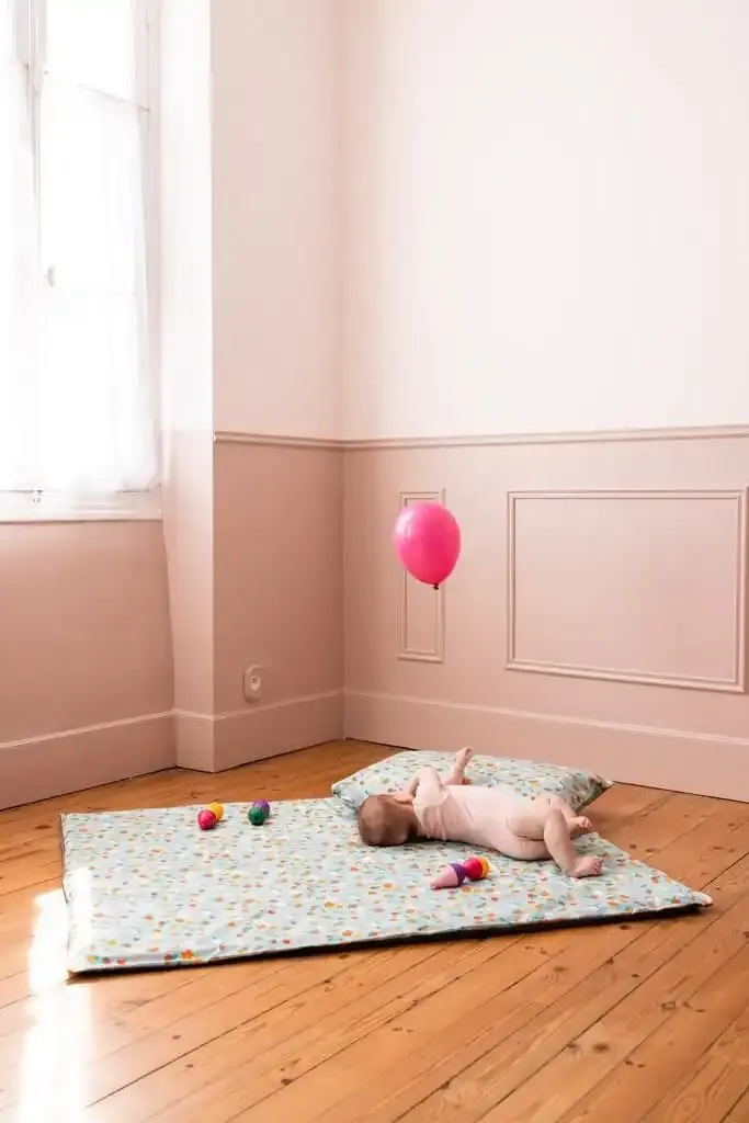 Un bébé allongé sur un tapis d'éveil clair à petits motifs, au milieu d'une chambre rose au parquet ancien, un ballon rose près du mur