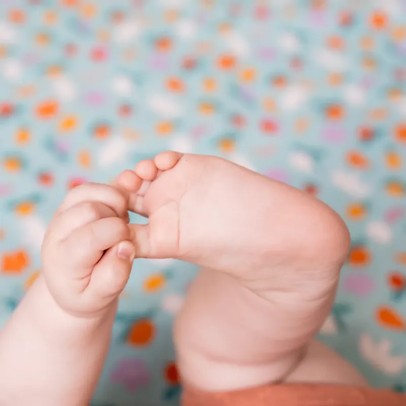 Gros plan sur les mains d'un bébé qui attrape son pied, sur fond de tapis d'éveil turquoise imprimé de fruits et de fleurs