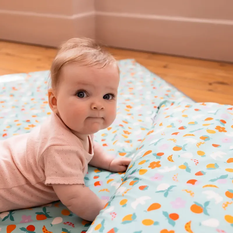 Un bébé en body rose appuyé sur les avant-bras sur un tapis d'éveil turquoise imprimé de fruits, qui regarde l'objectif