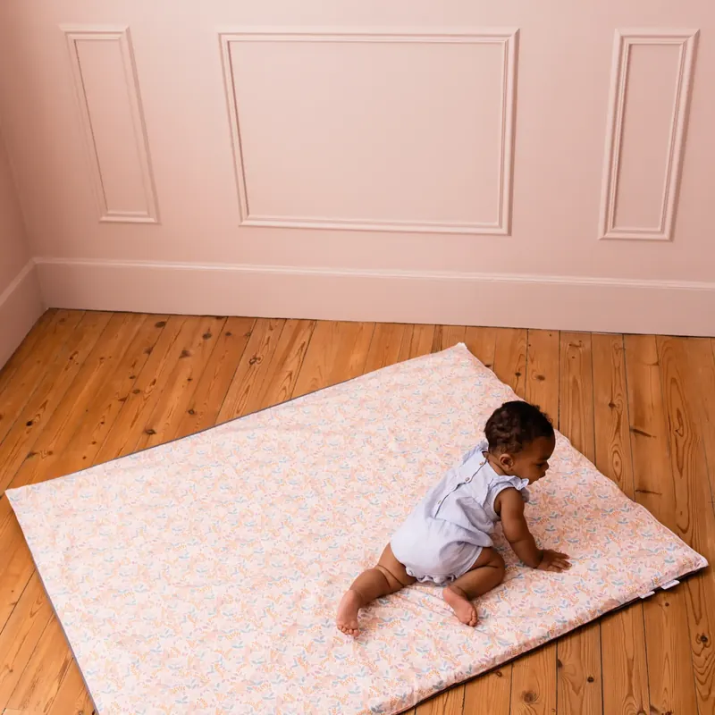 Un bébé en body blanc à quatre pattes sur un tapis d'éveil rose pâle à petites fleurs, au centre d'une pièce rose au parquet