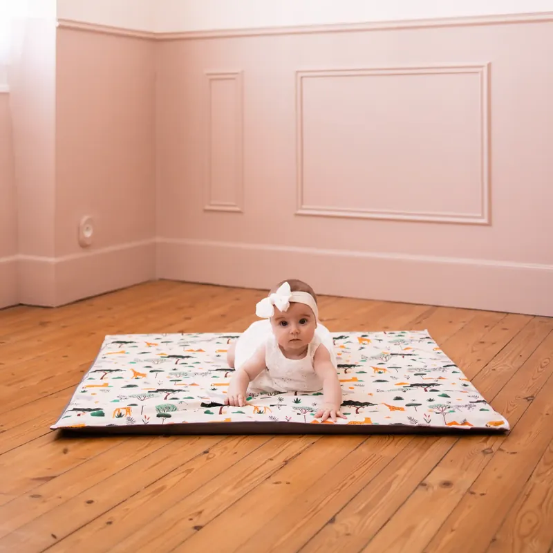 Un bébé en robe blanche et bandeau, à plat ventre sur un tapis d'éveil imprimé d'animaux de la savane, au milieu d'une pièce rose au parquet