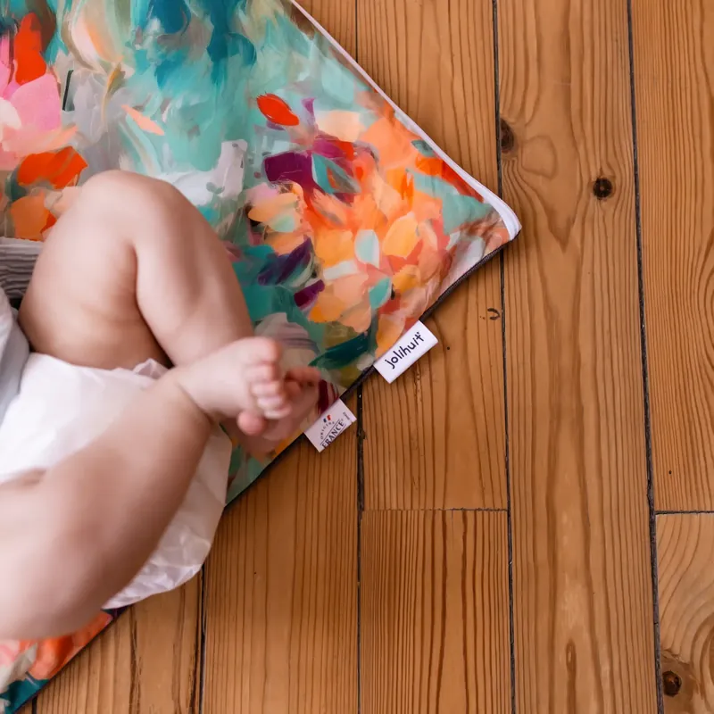 Les jambes d'un bébé en couche sur le coin d'un tapis d'éveil aux grandes taches de peinture turquoise et orange, posé sur un parquet
