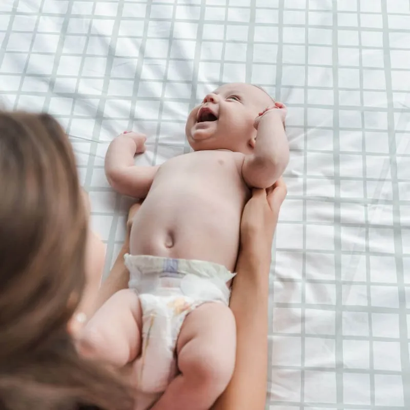 Un bébé en couche allongé sur le dos sur un tapis d'éveil à carreaux blanc et vert d'eau, qui rit aux éclats, tenu par sa maman