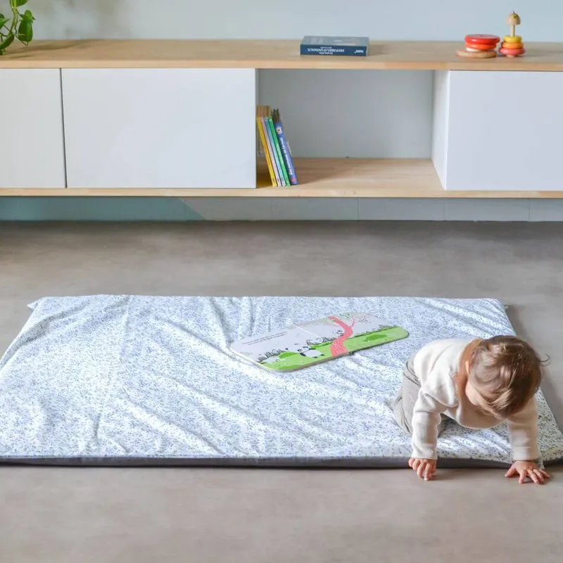 Un bébé qui rampe pour sortir d'un grand tapis d'éveil blanc à petits motifs bleus, un livre posé sur le tapis