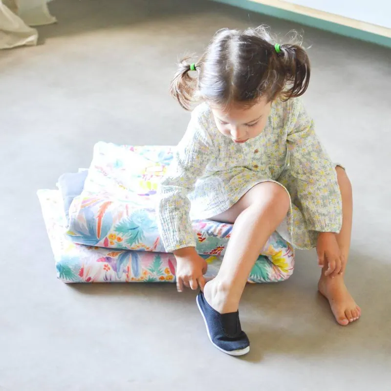Une petite fille à couettes assise sur un tapis d'éveil plié à imprimé jungle pastel, qui enfile sa chaussure
