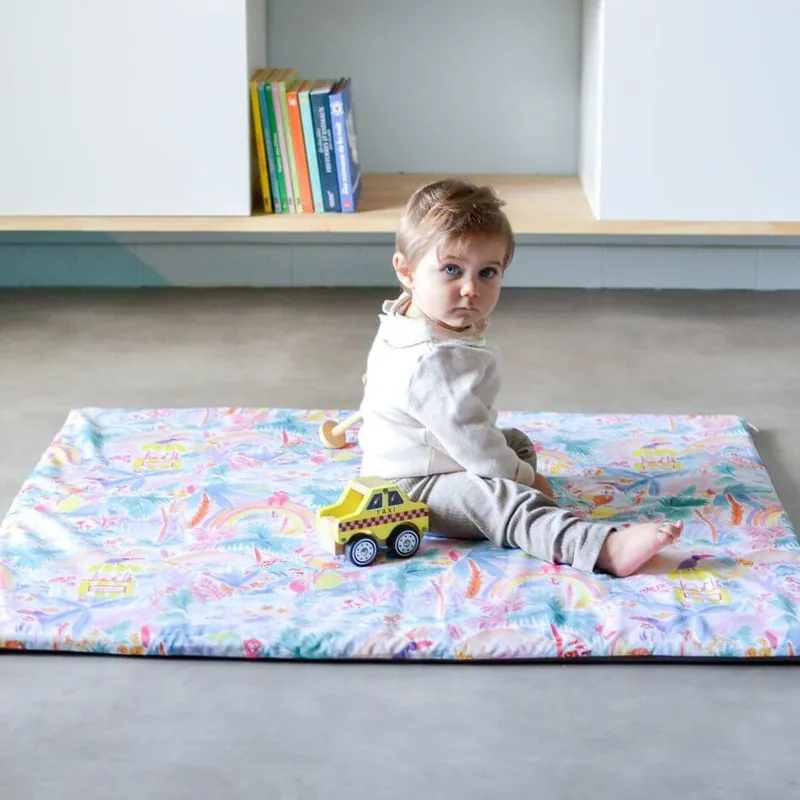 Un bébé assis sur un tapis d'éveil de taille standard à imprimé jungle pastel, une petite voiture jaune à côté de lui, se retourne vers l'objectif
