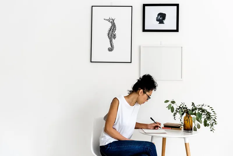 Une femme assise à un petit bureau blanc écrit sur une feuille, sous des cadres accrochés au mur