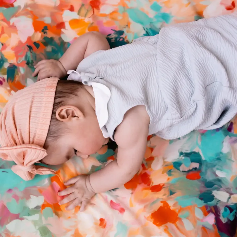 Un bébé coiffé d'un bandeau rose, allongé sur le ventre, joue sur un tapis d'éveil imprimé de grandes fleurs orange et bleues