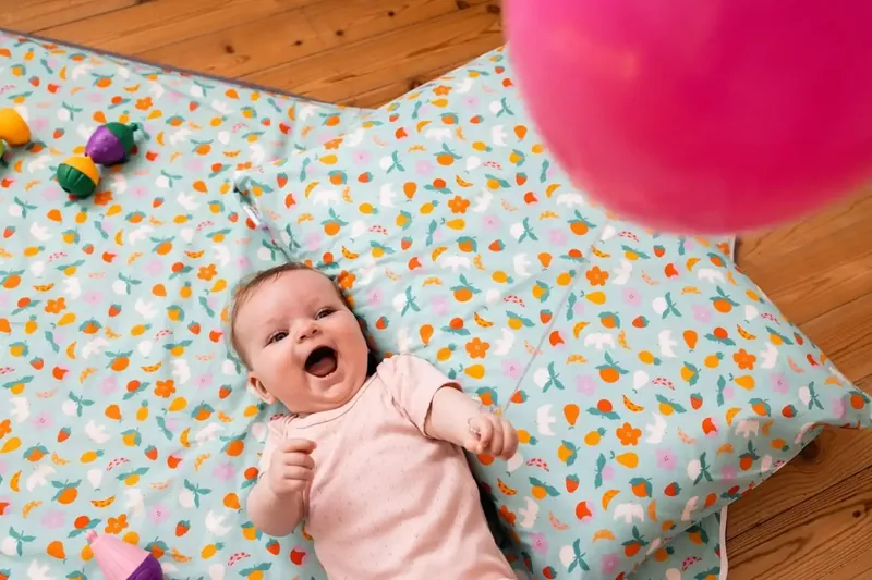 Un bébé en body rose, allongé sur le dos, rit sur un tapis d'éveil imprimé de fruits posé sur un parquet, près d'un ballon rose