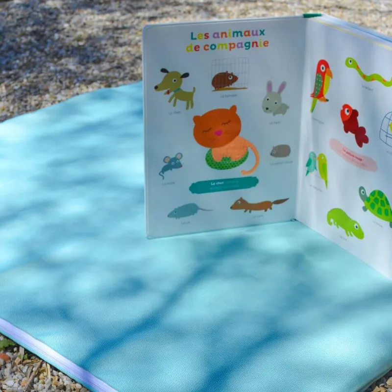 Un imagier cartonné « Les animaux de compagnie » ouvert et posé debout sur un tapis d'éveil turquoise, dehors sur des graviers