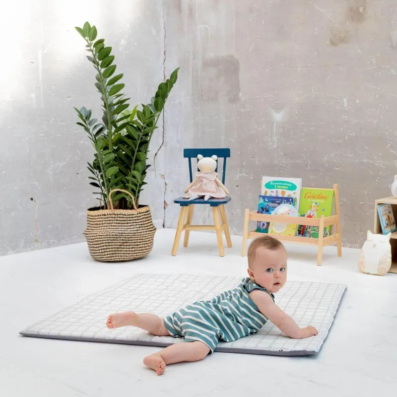 Un bébé en barboteuse rayée verte, allongé sur le ventre sur un tapis d'éveil à carreaux gris, devant une petite chaise et une bibliothèque d'enfant