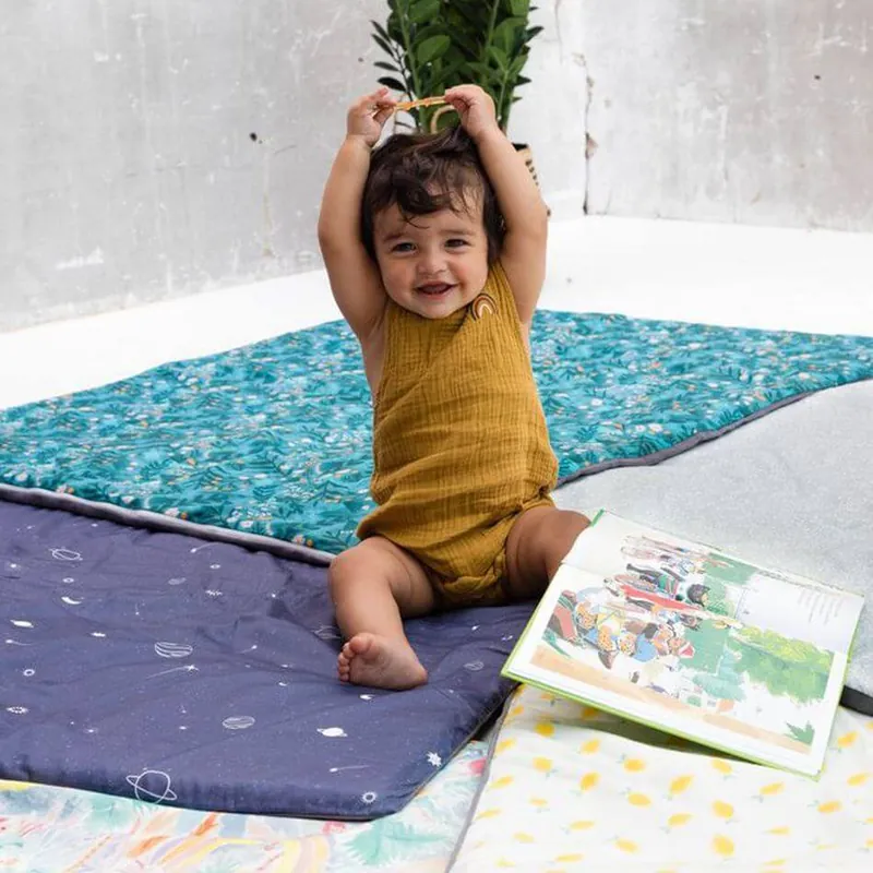 Une petite fille en barboteuse moutarde, assise sur des tapis d'éveil dépliés, les mains sur la tête, à côté d'un livre ouvert