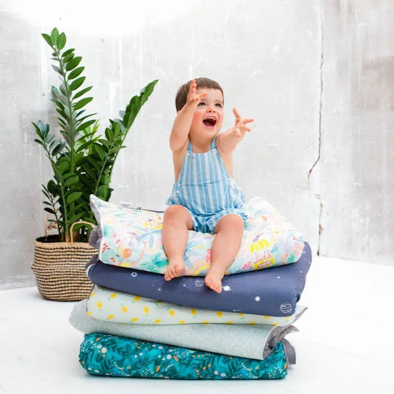 Un petit garçon en salopette rayée bleue, assis en haut d'une pile de tapis d'éveil pliés, lève les bras en riant