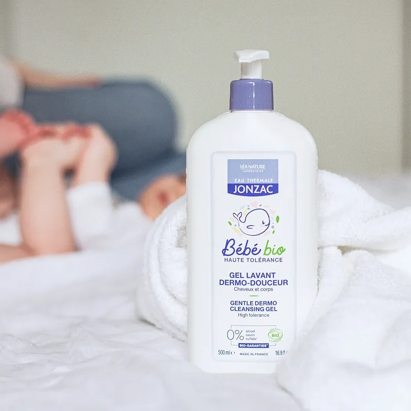 Un flacon pompe de gel lavant dermo-douceur Bébé bio Eau Thermale Jonzac posé sur un drap blanc, devant un bébé flou