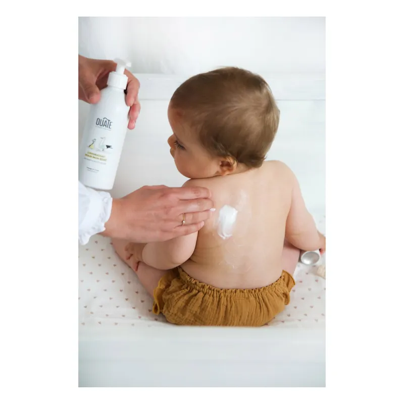 Un adulte applique une crème Ouate sur le dos d'un bébé assis en culotte moutarde, un flacon à la main