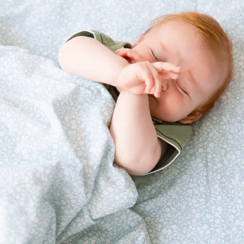 Un bébé roux, allongé sur un tapis d'éveil bleu ciel à petites fleurs blanches, se frotte les yeux