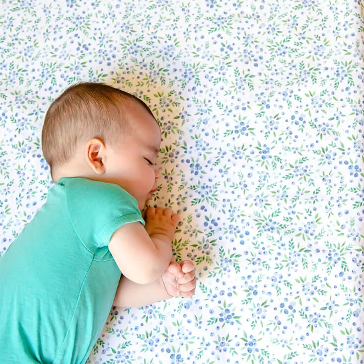 Un bébé en body turquoise dort sur le côté sur un tapis d'éveil blanc à petites fleurs bleues